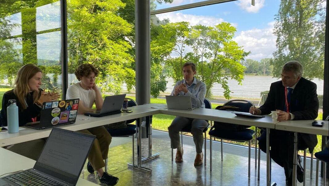 Cari Anna og Adam sitter i et møterom med to andre delegater/observatører foran et stort vindu med grønne trær og vann i bakgrunnen.